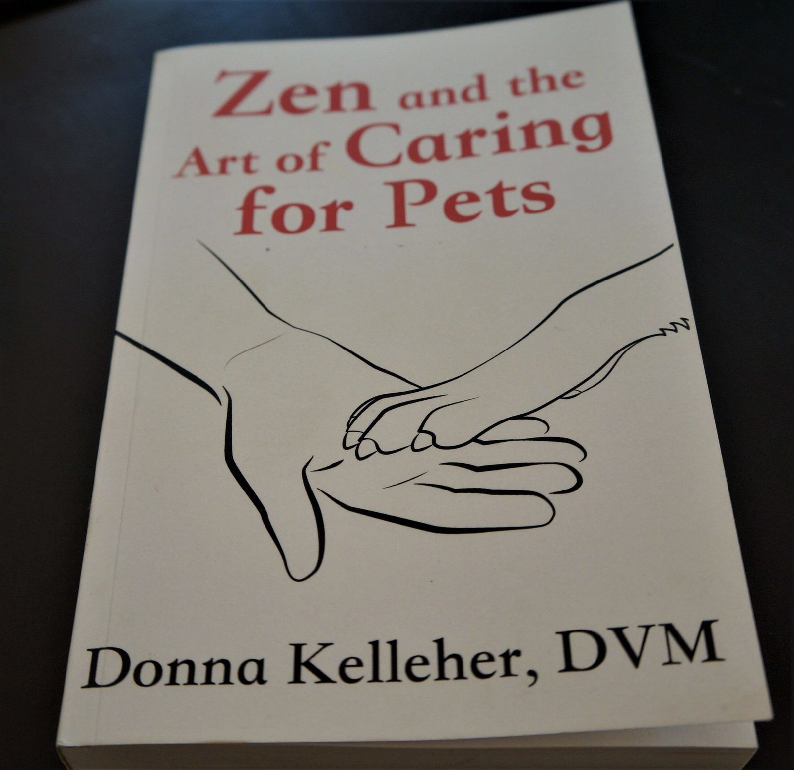 Zen and the art of caring for pets book cover - white paper with red test with black line drawing of a person's hand with a pet's paw in it